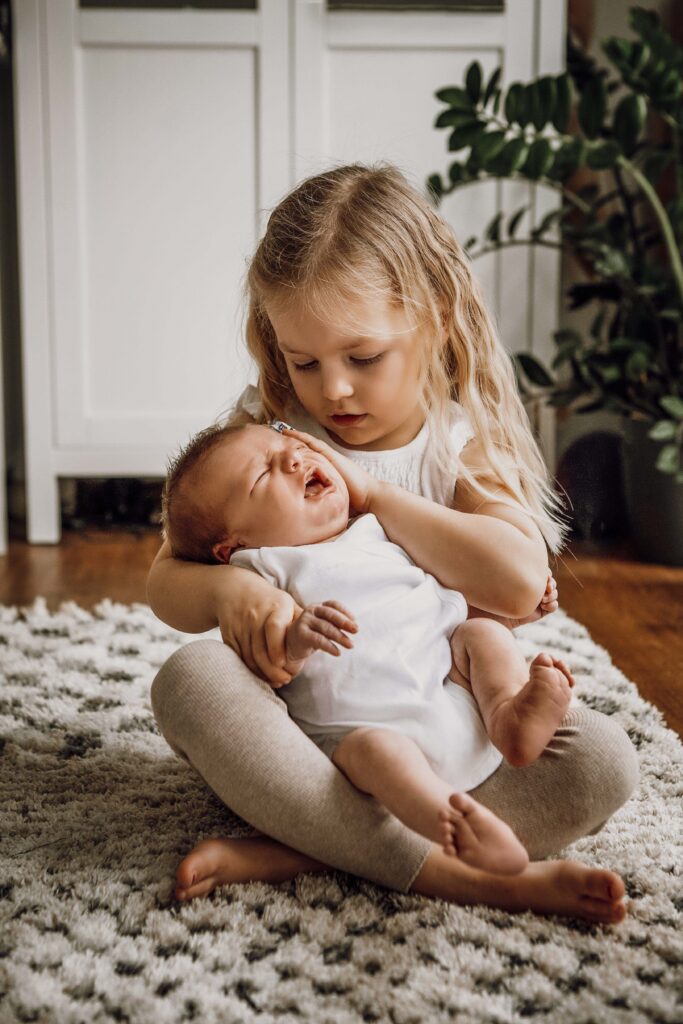 Große Schwester sitz mit dem Baby auf dem bode und streichelt ihn während er weint.