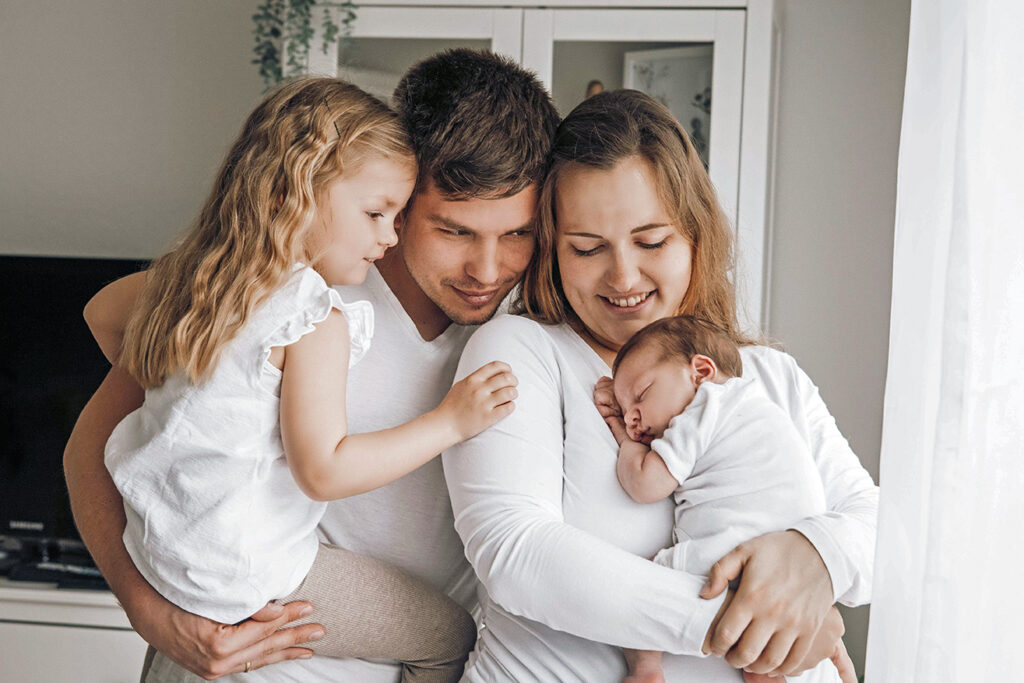 Familie schaut baby an während alle zusammen vor dem Fenster stehen und jeder ein Kind auf dem arm hat. 