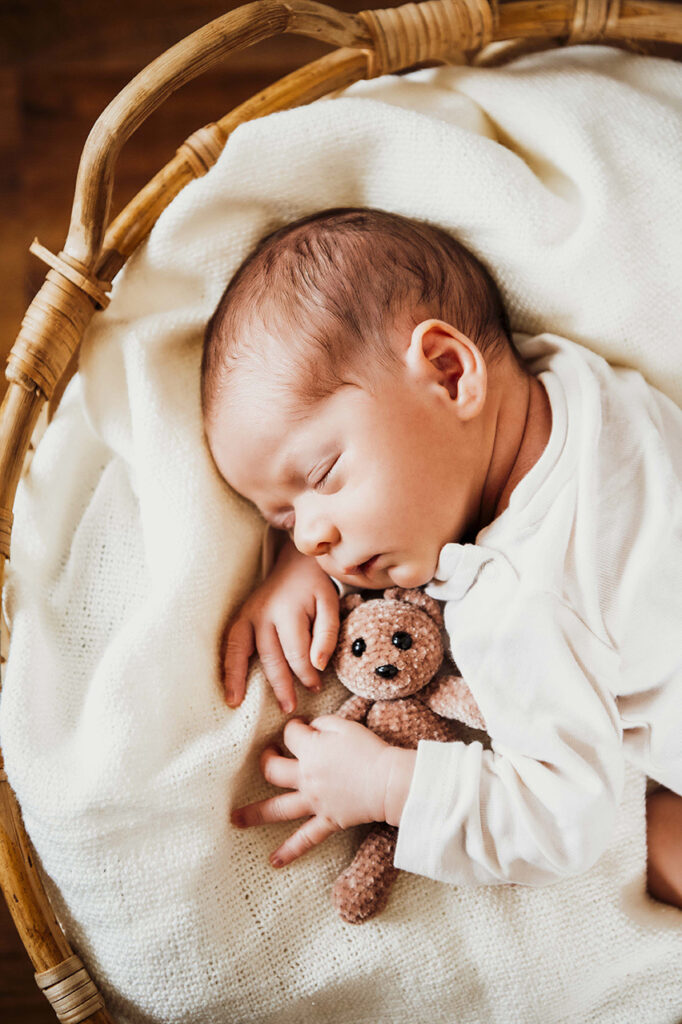 Baby liegt im Body im Körbchen und hält einen kleinen Bär.