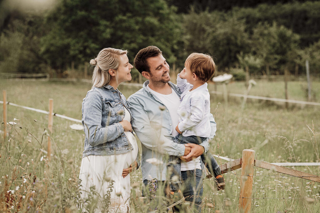 eine Kleine Familie lacht miteinander und steht in einer hohen wiese. der Papa hat den Sohn auf den arm.
