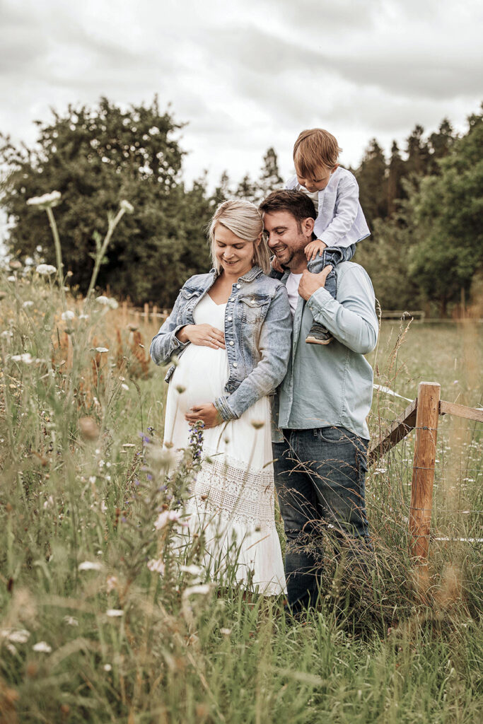 Familie steht in der hohen wiese. der große Bruder sitzt auf Papas Schultern und alle schauen auf den Babybauch.