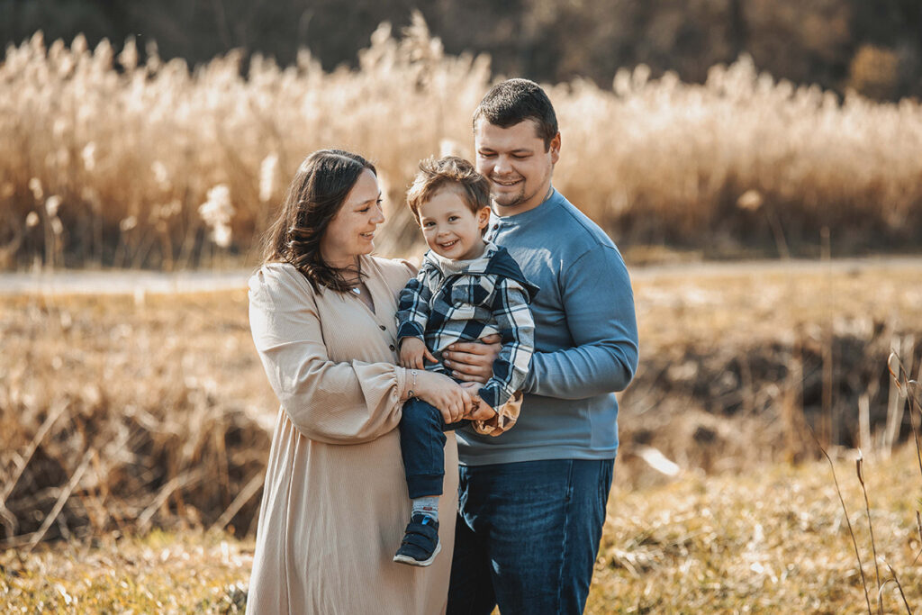 Familien Bild Schwangere Mama hält den Sohn auf dem arm und lacht ihn an, der Papa lacht ihn auch an.