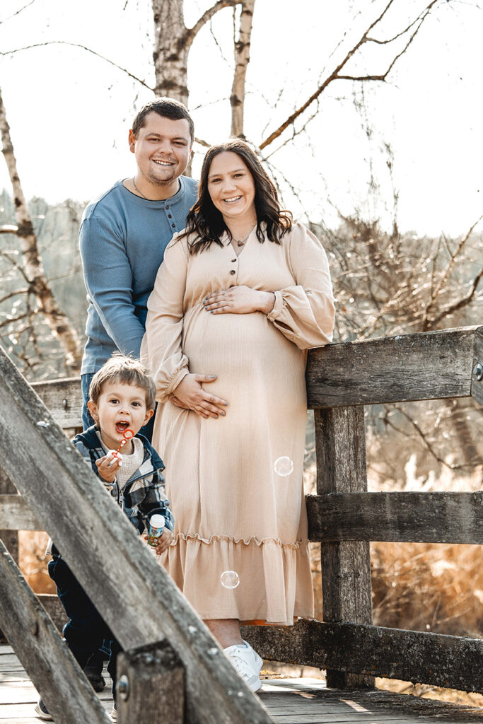 Familie steht auf einer alten Holzbrücke, die mama hält den Babybauch. der Sohn macht im Vordergrund Seifenblasen.
