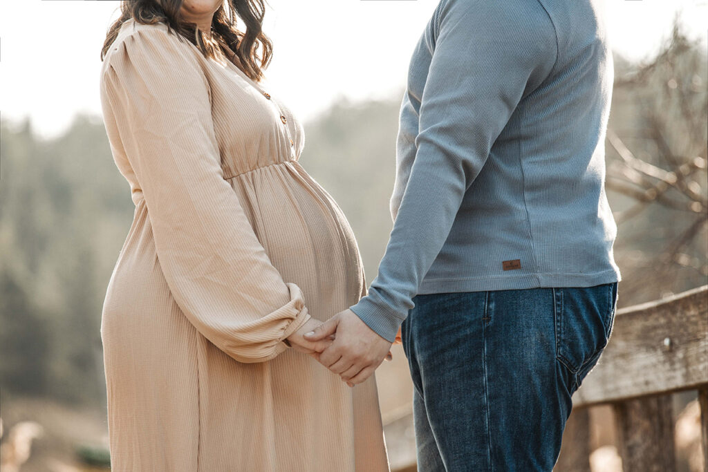 Zeigt  ein Babybauch als Nahaufnahme auf einer Brücke. die Eltern halten sich Hand in Hand.