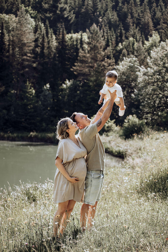 Junge Familie steht am See und der Papa wirft den Sohn hoch. während die Mama ihren Babybauch hält.