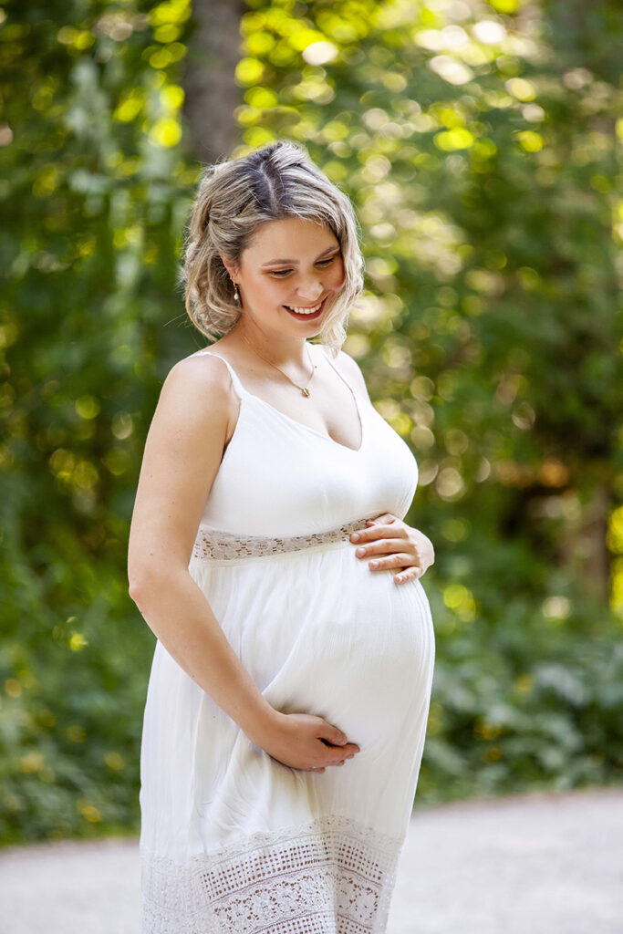 Schwangere Frau in einem Weißen Kleid, steht im Wald und schaut auf den Bauch.