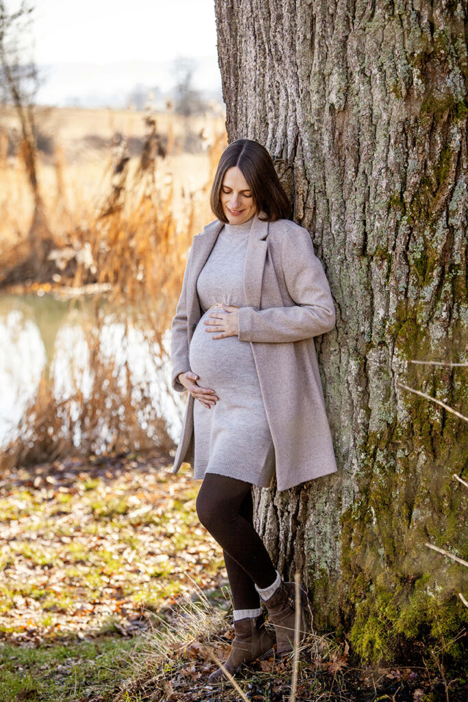 Junge Frau in Herbstkleidung schaut auf den Bauch während sie sich an den Baum lehnt.