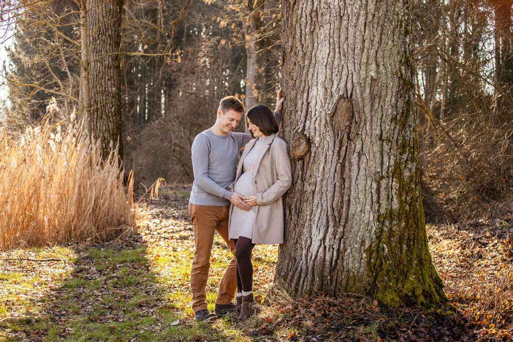 werden Eltern stehen im Herbst an einem Baum gelehnt und schauen erwartungsvoll auf den Babybauch.