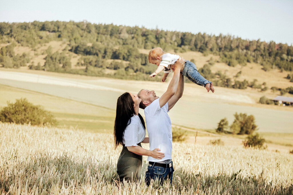 Papa hebt den Jungen über den Kopf und macht Quatsch. Die Mama lacht von Hinten den Jungen an.