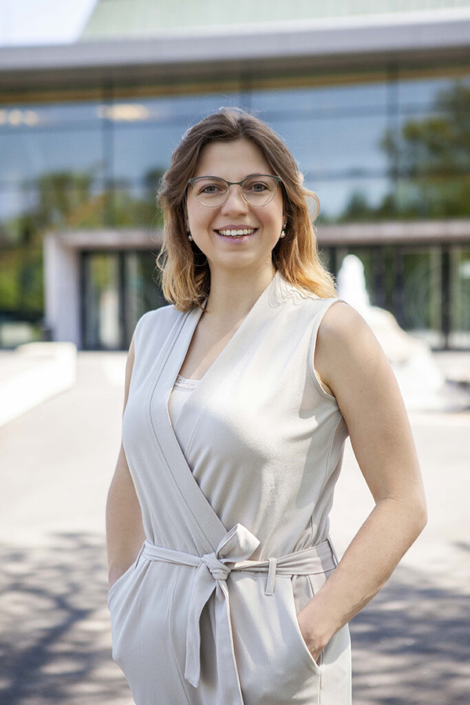 Nahaufnahme einer Frau Braunes Haar und Brille lachend vor der Stadthalle in Balingen mit Handen in der Hose. im Hintergrund ist der Springbrunnen unscharf.