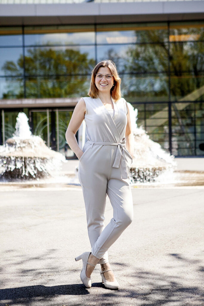 Nahaufnahme einer Frau Braunes Haar und Brille lachend vor der Stadthalle in Balingen mit Handen in der Hose. im Hintergrund ist der Springbrunnen unscharf.