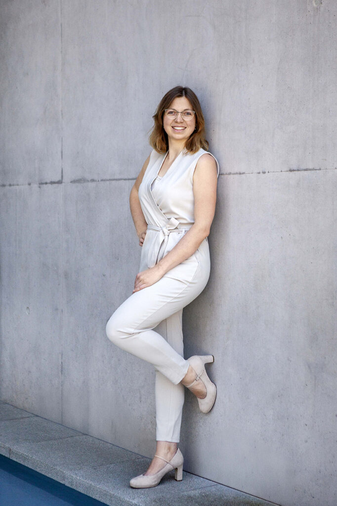 Frau steht in Balingen Stadthalle an der Beton Wand im Hosenanzug mit Handen in der Hose und Bein an der wand.