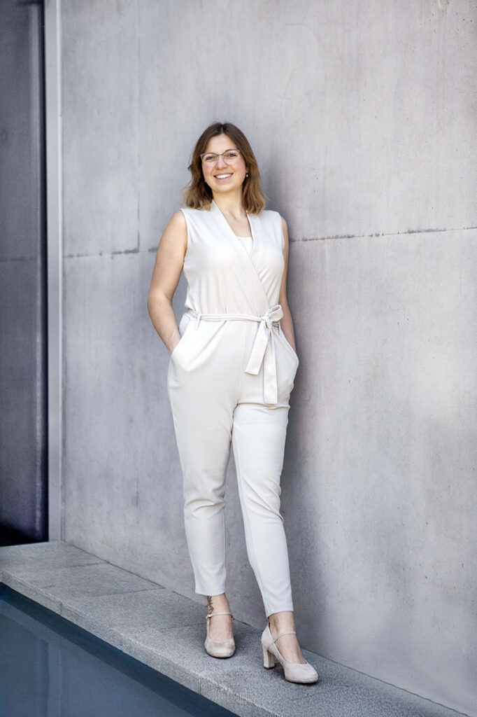 Frau steht in Balingen Stadthalle an der Beton Wand im Hosenanzug mit Handen in der Hose.