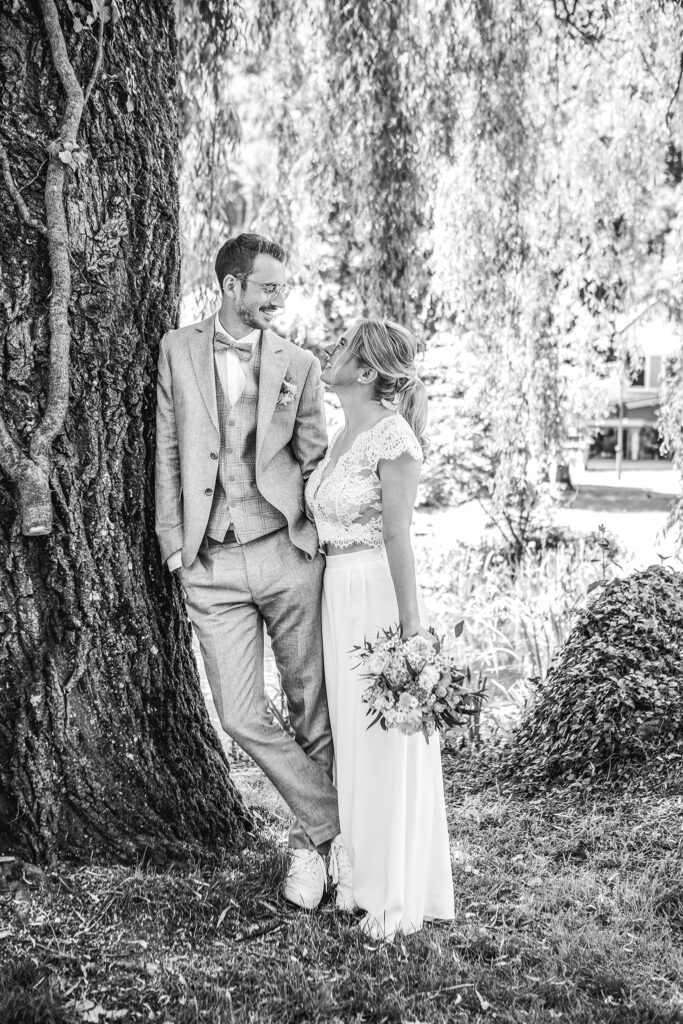Brautpaar Steht Hand in Hand im Bad Imnau im Park und lachen miteinander während der Mann am Baum lehnt. Bild Schwarzweiß.