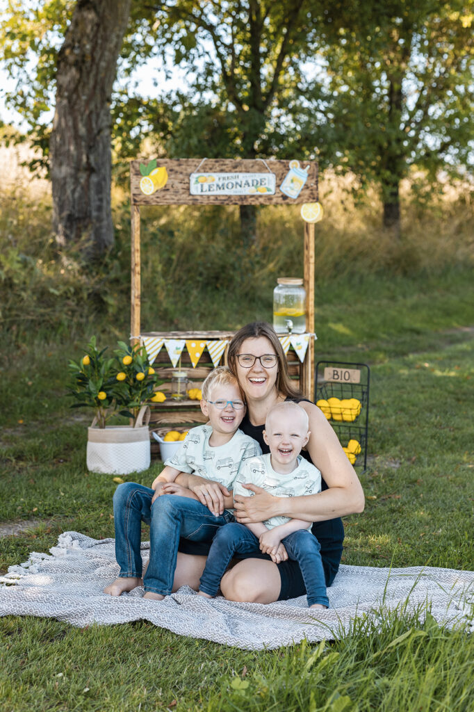 Mama sitz mit Zwei Jungs  auf einem Teppich in der Wiese im Schneidersitz und lacht. Im Hintergrund ist ein Limonaden Stand Empfingen.